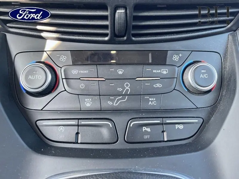 Vue rapprochée de la console centrale noire du Ford Kuga blanc, montrant les commandes de climatisation et d’aide au stationnement.