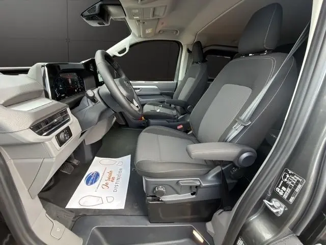 Vue intérieure côté conducteur du Volkswagen Transporter gris foncé 2026, sièges tissu avec accoudoirs et tableau de bord moderne.