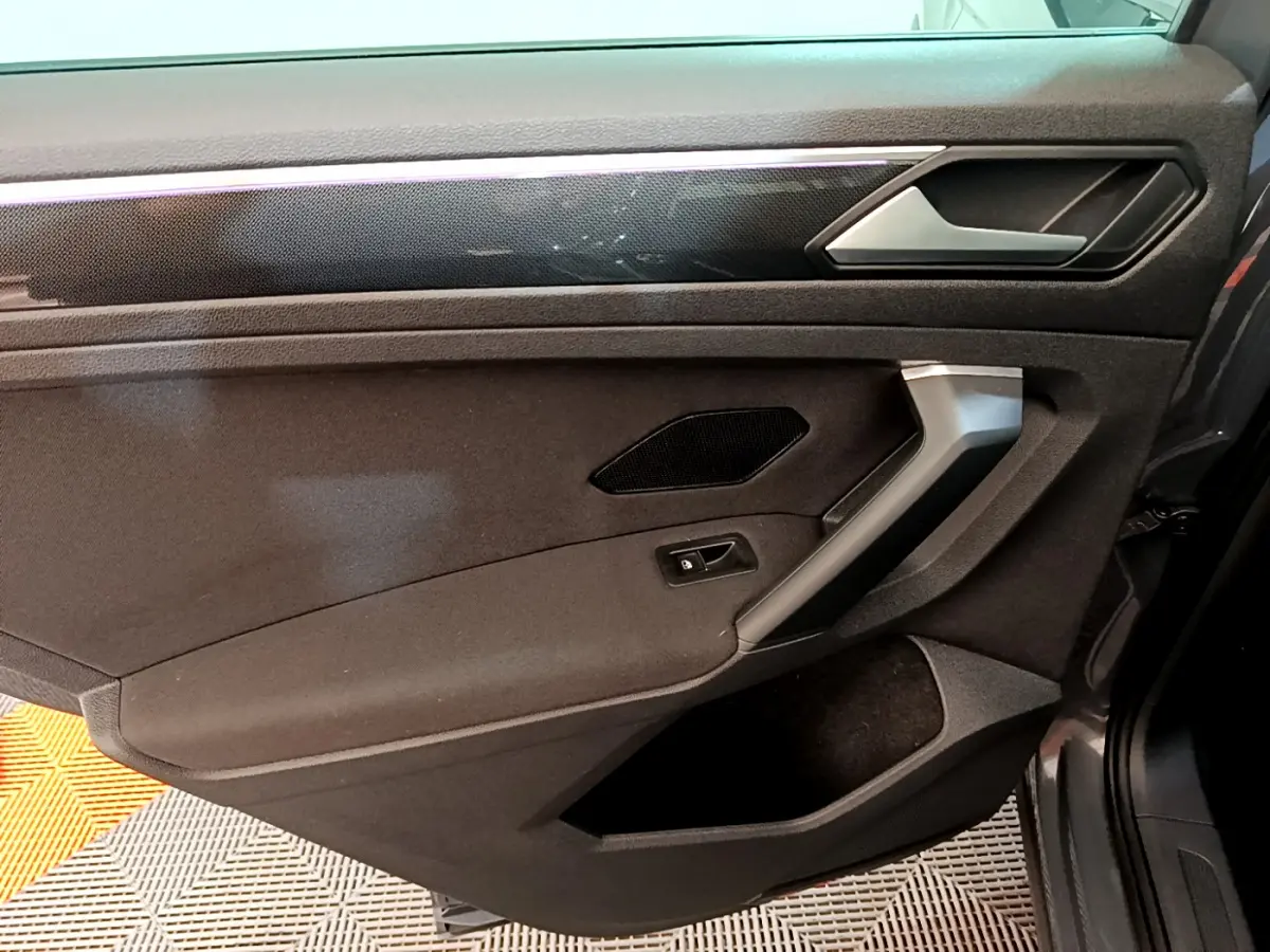Vue intérieure côté droit de la porte du Volkswagen Tiguan gris clair, avec poignée argentée et panneau noir texturé.