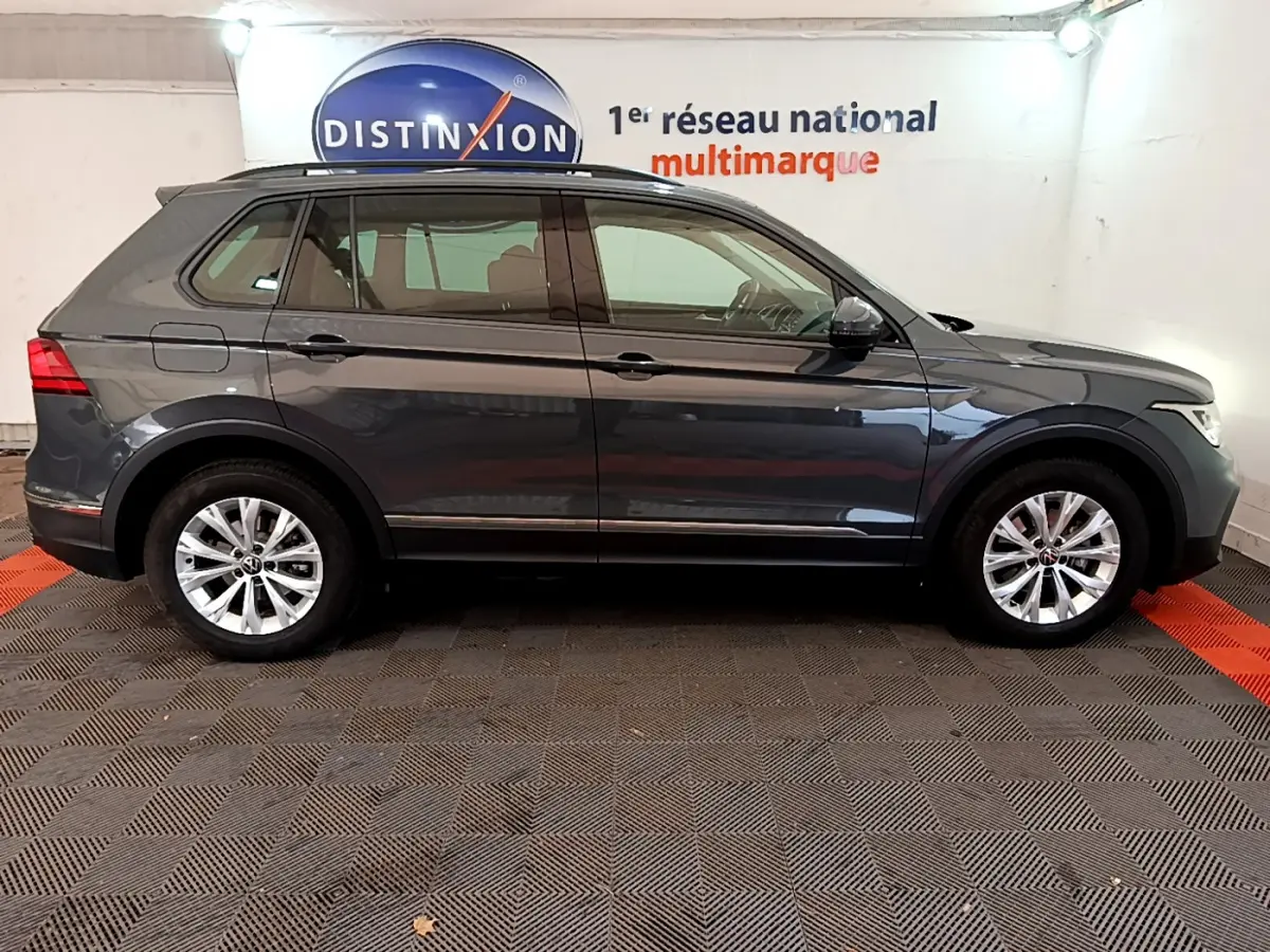 Volkswagen Tiguan gris clair vu de profil côté gauche dans un showroom avec logo Distinxion en arrière-plan.