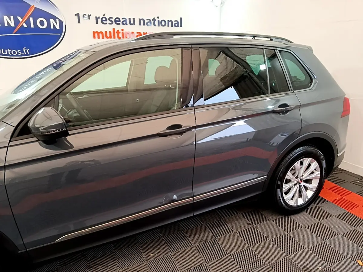 Vue latérale côté gauche d'un Volkswagen Tiguan gris clair 2021, avec jantes alliage et barres de toit noires visibles.