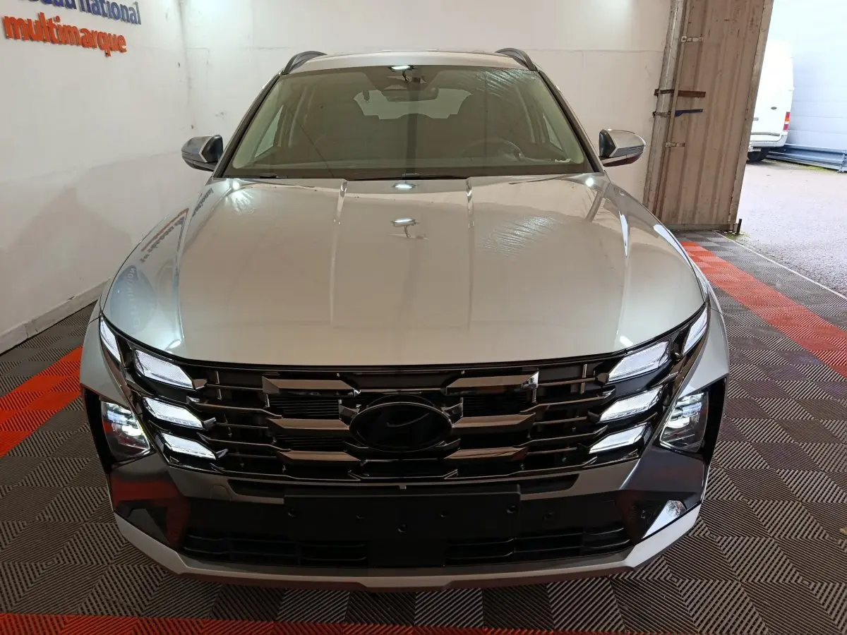 Vue avant d’un Hyundai Tucson gris clair 2025 avec calandre noire et feux LED distinctifs, en intérieur showroom.