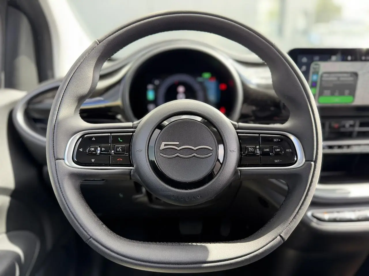 Volant noir Soft-Touch de la FIAT 500 électrique 2024 vu de face avec commandes intégrées et tableau de bord flou.