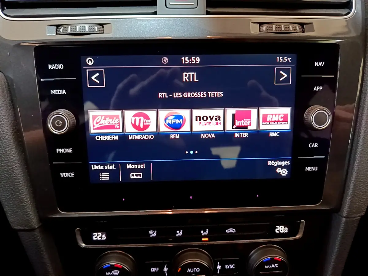 Vue rapprochée de l’écran tactile central de la Volkswagen Golf noire, affichant les stations radio et commandes climatisation.