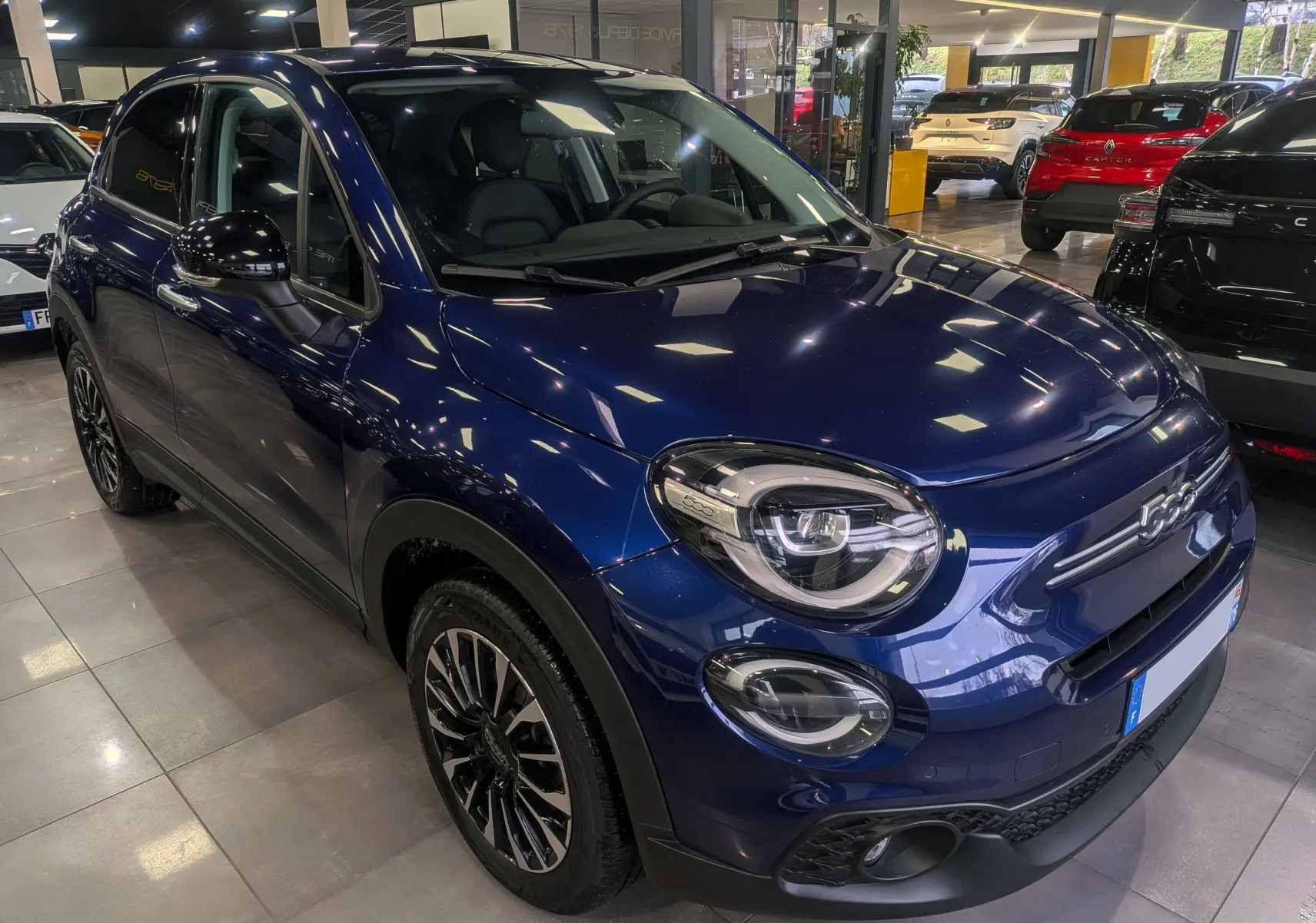 Vue 3/4 avant gauche d'une FIAT 500X 2024 bleu Venezia avec jantes noires et phares LED en showroom.