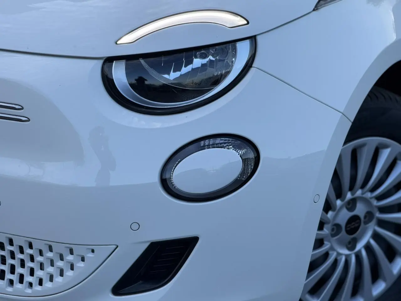 Gros plan sur le phare avant gauche blanc Artic de la FIAT 500 électrique 2024 avec jante alliage visible.