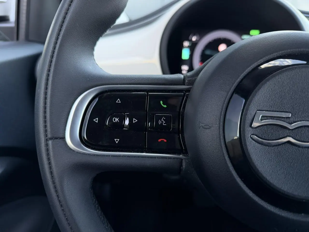 Gros plan sur la partie gauche du volant noir de la Fiat 500 électrique 2024 avec commandes intégrées et logo central.