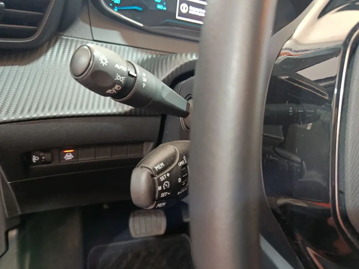 Gros plan sur la colonne de direction et commandes au volant d'une Peugeot 208 électrique gris clair, tableau de bord partiellement visible.
