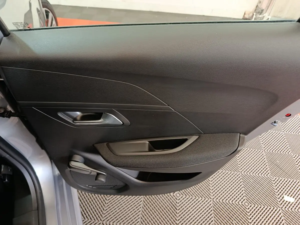 Intérieur côté gauche de la porte avant d'une Peugeot 208 électrique gris clair, avec poignée et commande de vitre manuelle.