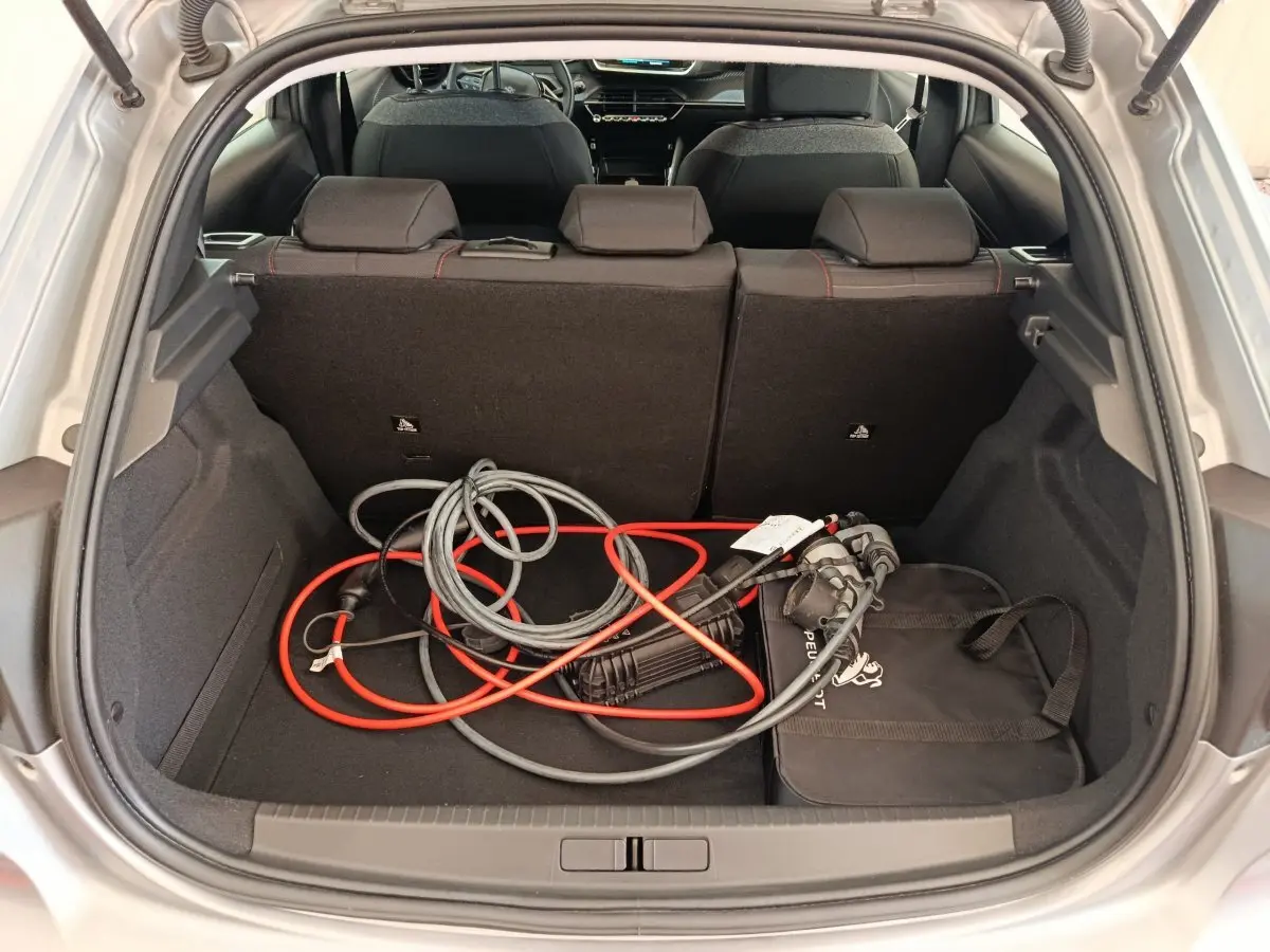 Coffre ouvert d'une Peugeot 208 électrique gris clair, montrant câbles de recharge et banquette arrière rabattable.
