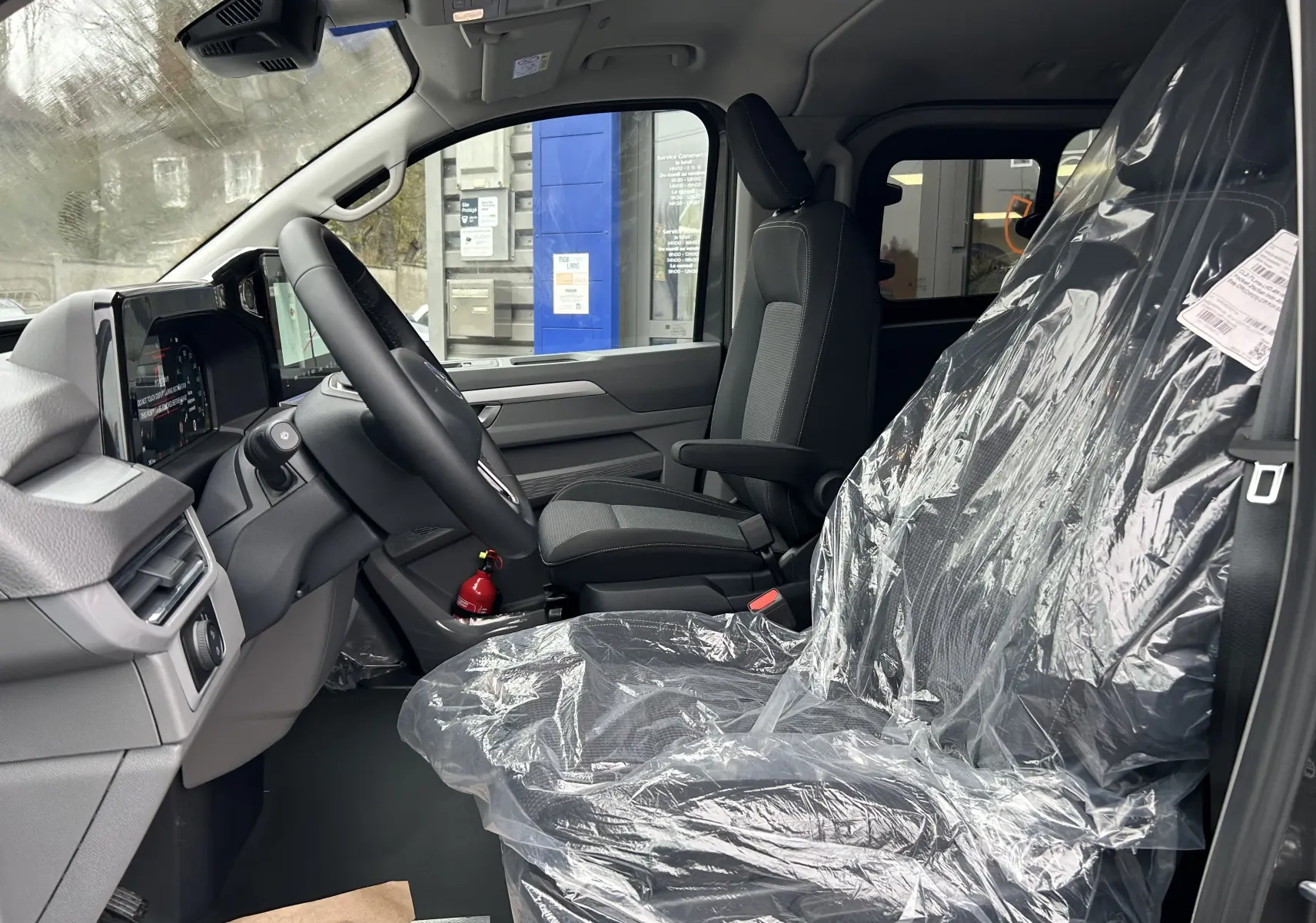 Vue intérieure côté conducteur du Volkswagen Transporter T7 ProCab, sièges noirs avec protection plastique, tableau de bord moderne.