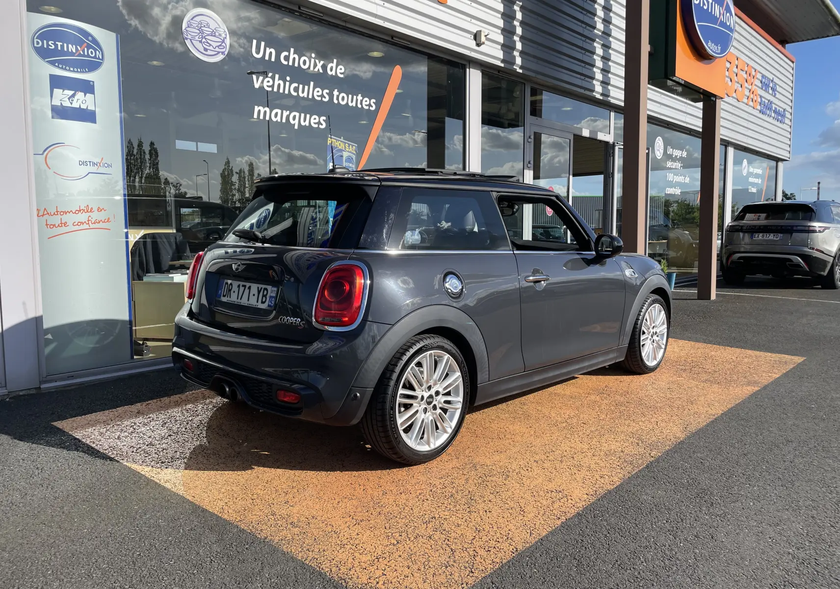 Vue 3/4 arrière droite d'une MINI F56 Cooper S gris Thundergrey avec jantes 17 pouces et toit noir devant un showroom.