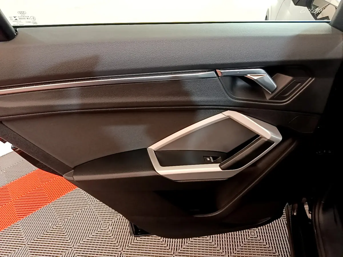 Vue intérieure côté gauche de la porte avant noire d’un Audi Q3 35 TDI 2021 avec poignée argentée et commandes fenêtres.