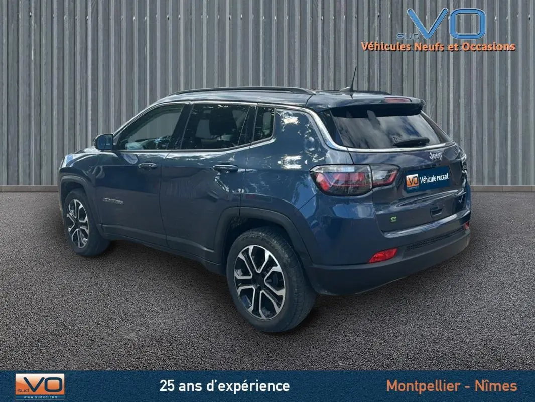 Vue 3/4 arrière droite d'un Jeep Compass bleu 2023 avec barres de toit et jantes 18 pouces, stationné devant un mur métallique.