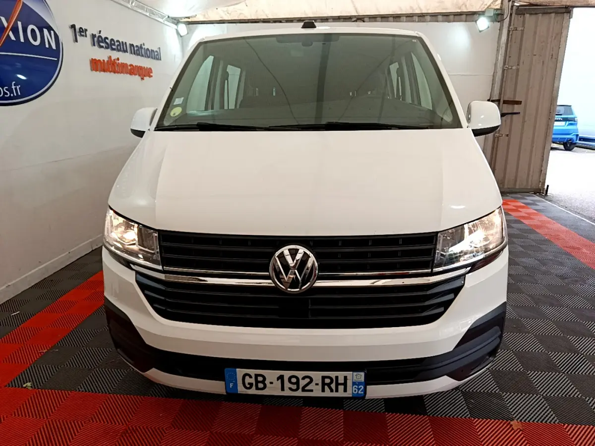 Vue avant d’un Volkswagen Transporter blanc 2021 avec calandre noire et phares allumés dans un garage.