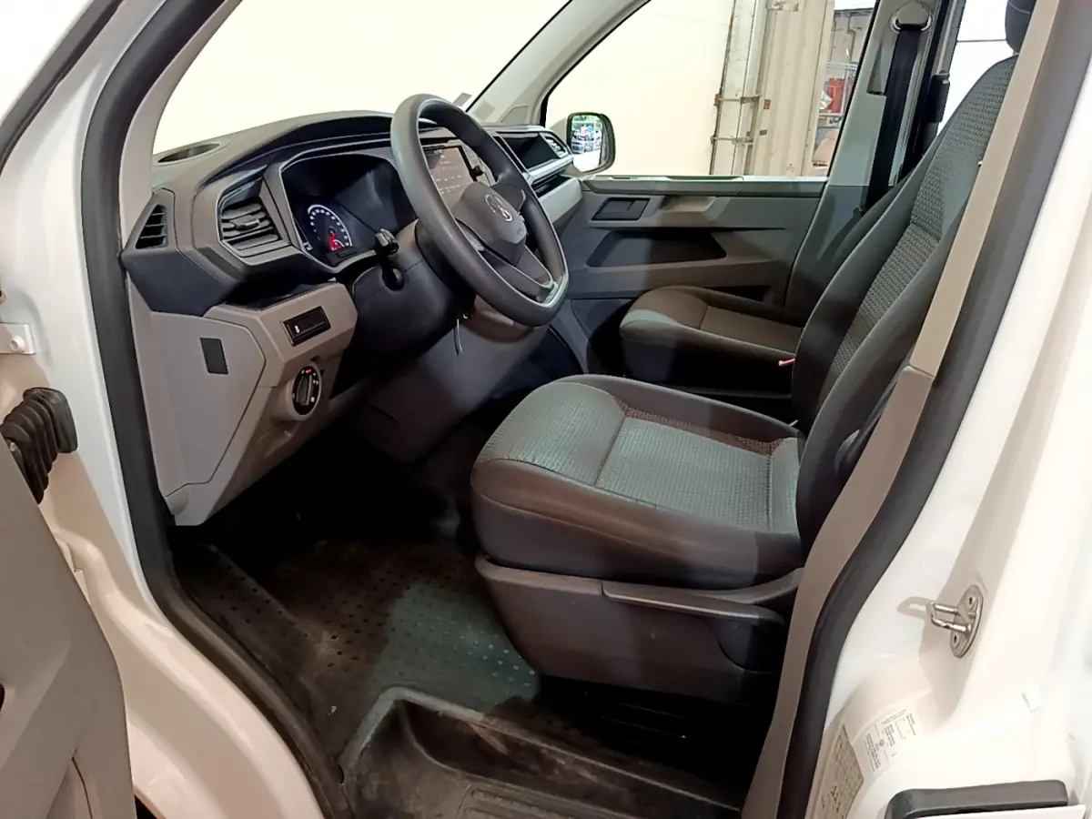 Vue intérieure côté conducteur du Volkswagen Transporter blanc, montrant le tableau de bord et les sièges avant gris.