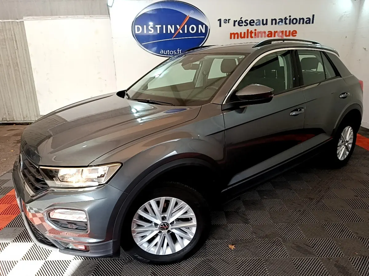 Volkswagen T-Roc gris foncé en 3/4 avant droit, avec jantes alliage et feux allumés en intérieur showroom.