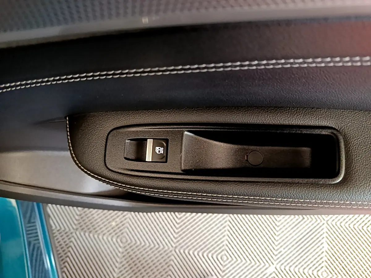 Gros plan sur la poignée intérieure et commande de vitre du côté gauche, avec surpiqûres blanches et teinte bleu visible à l’extérieur.