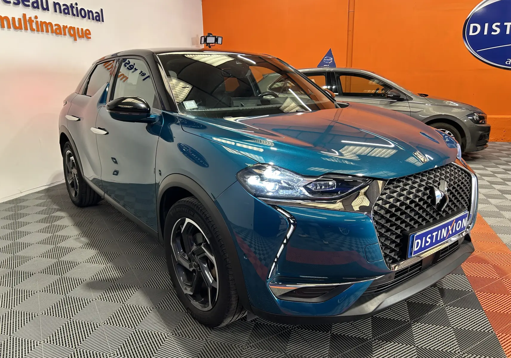 DS3 Crossback Bleu Millenium en 3/4 avant droit, avec calandre distinctive et phares allumés dans un showroom.