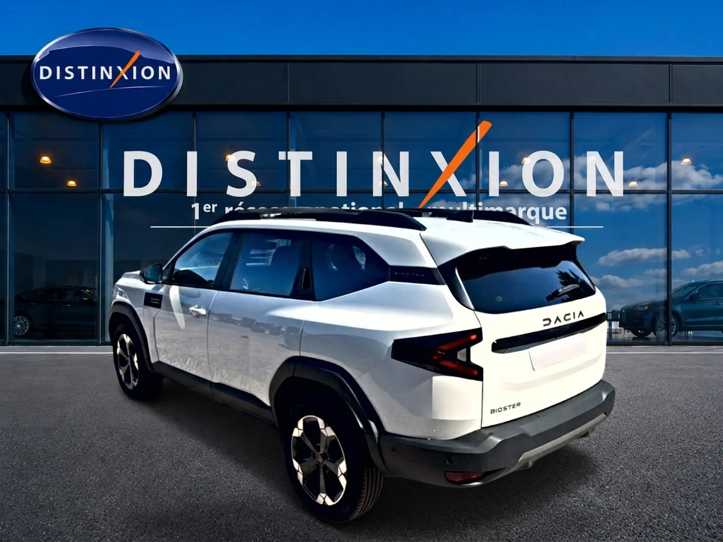 Vue 3/4 arrière droite du Dacia Bigster blanc avec barres de toit et jantes alu devant un showroom Distinxion.