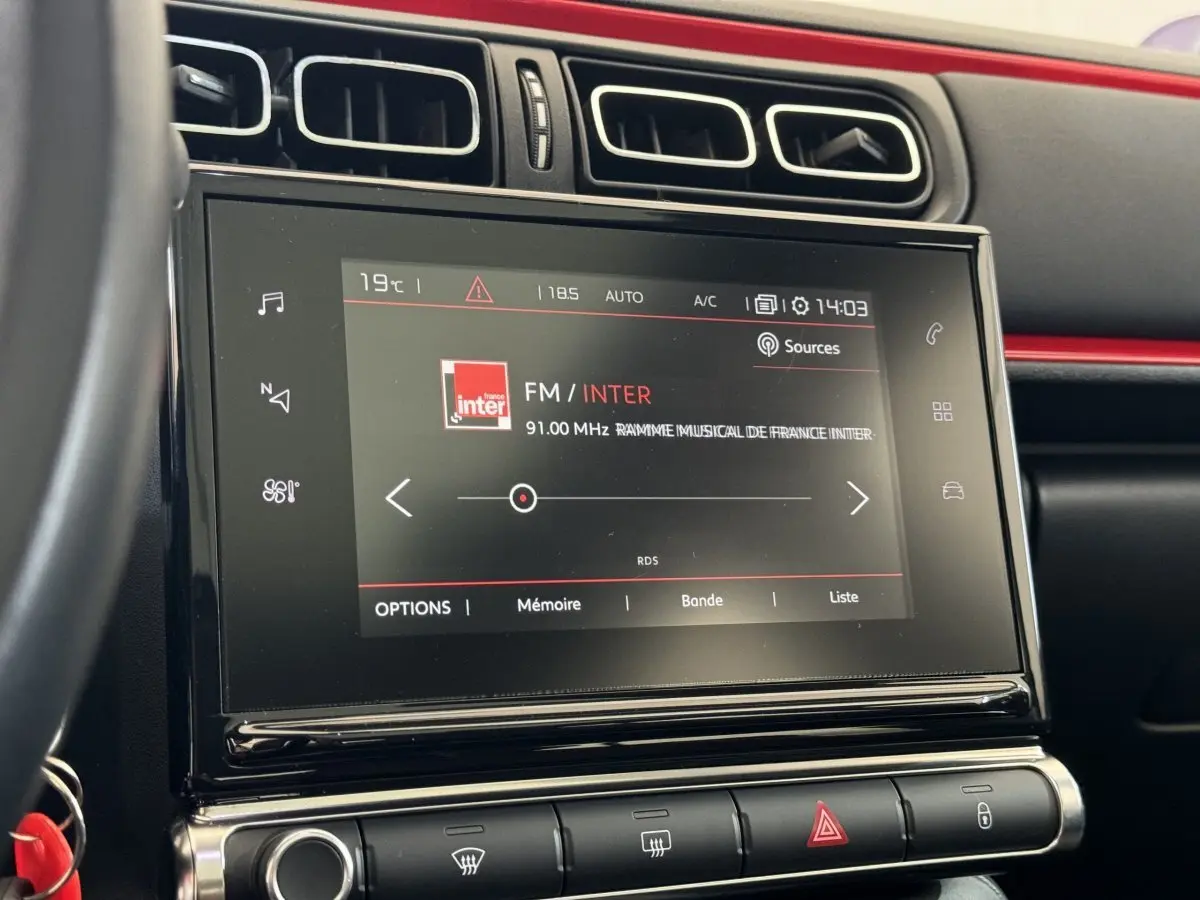 Écran tactile central de la Citroën C3 noire, vue rapprochée du tableau de bord avec commandes climatisation et radio FM affichée.