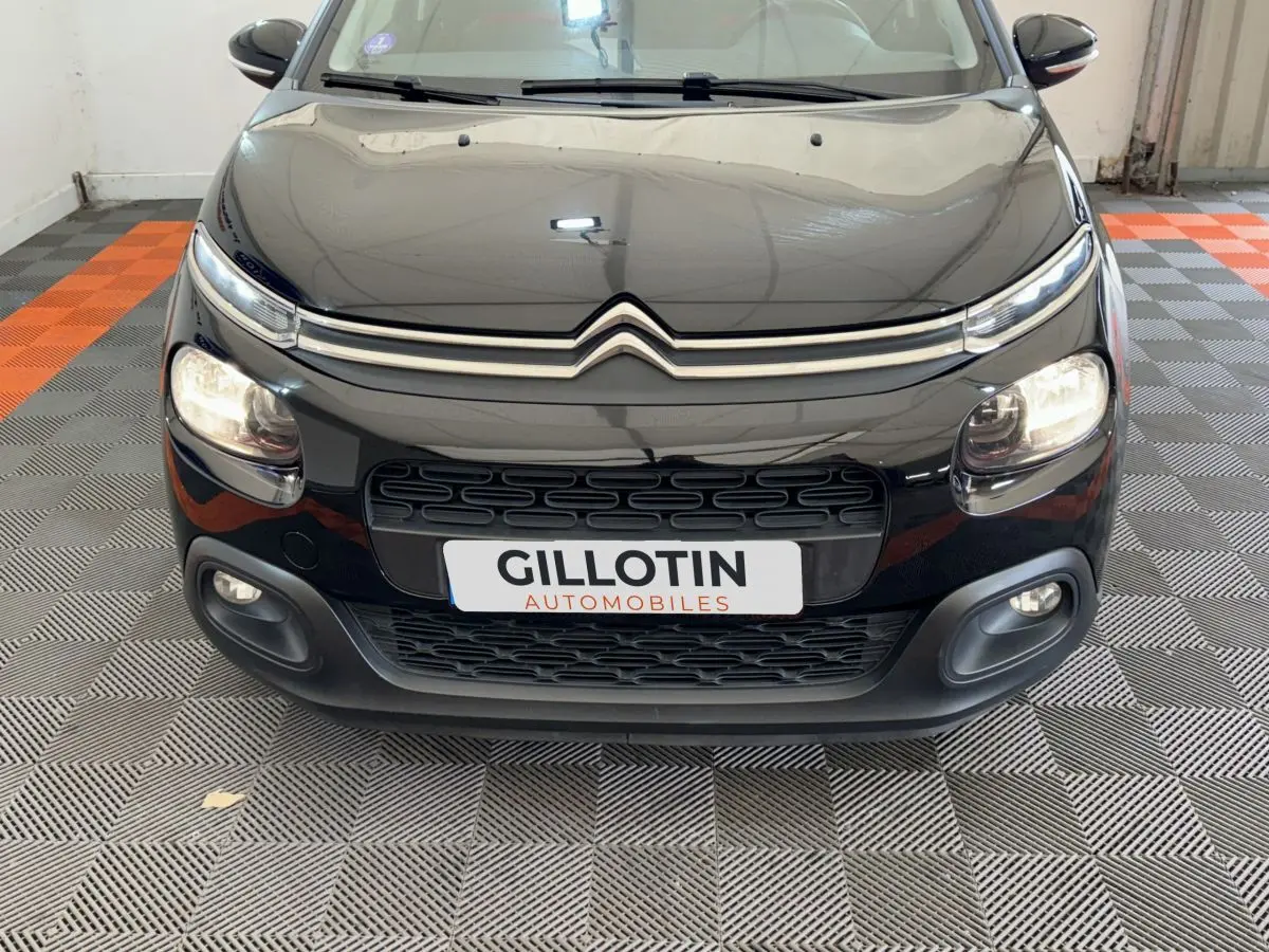 Vue frontale rapprochée d'une Citroën C3 noire avec phares allumés et calandre distinctive en intérieur showroom.