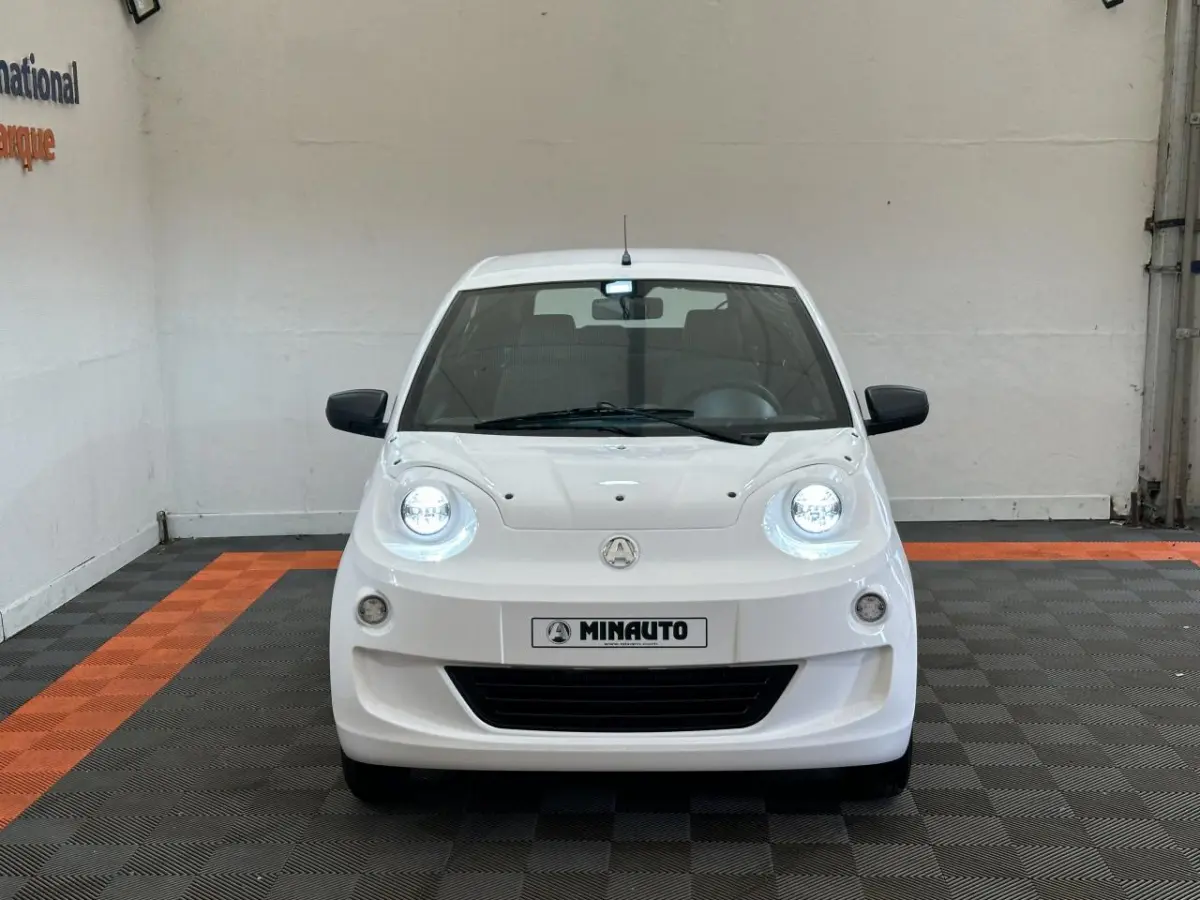 Vue frontale d'une AIXAM MINAUTO E ACCESS blanche 2025 avec phares allumés et rétroviseurs noirs côté conducteur.