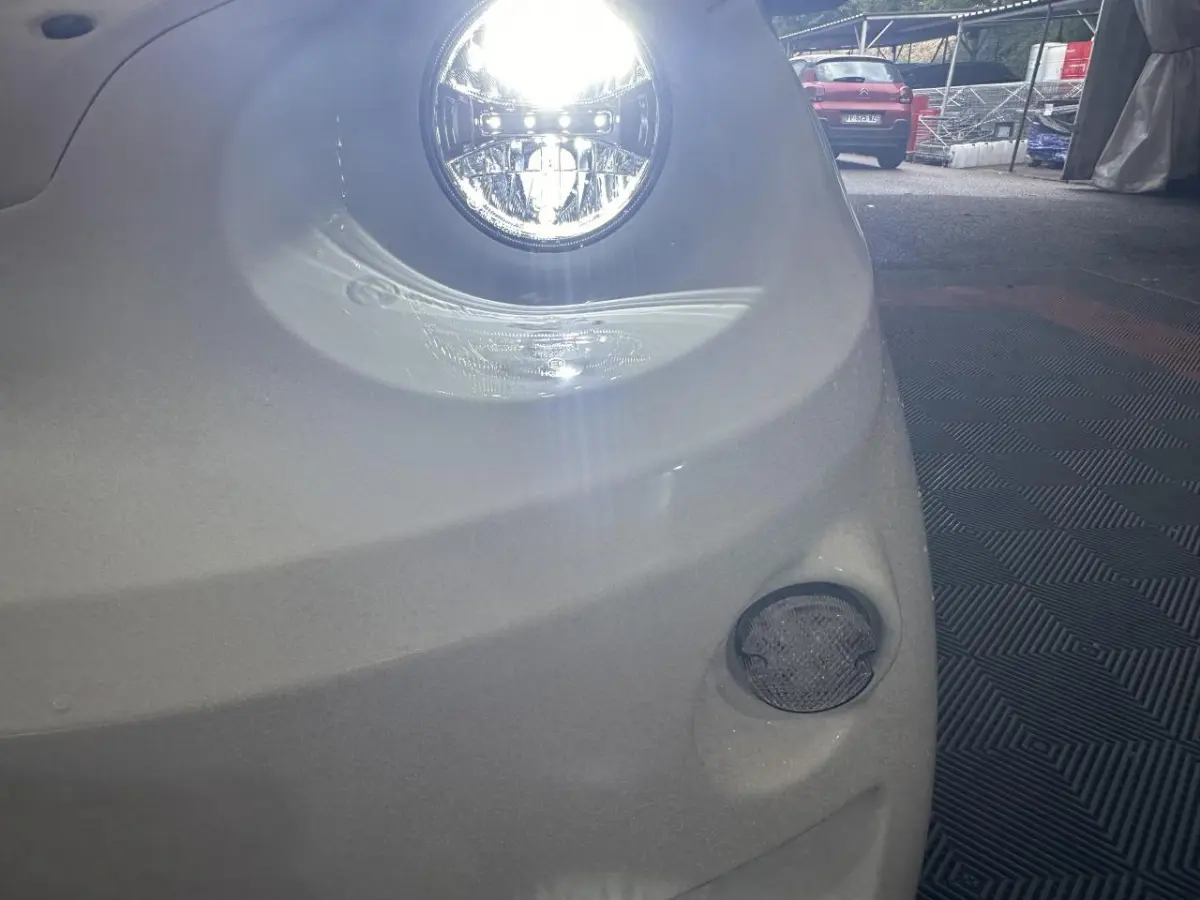 Gros plan sur le phare avant droit allumé d'une Aixam Minauto E Access blanche 2025 avec feu antibrouillard visible.