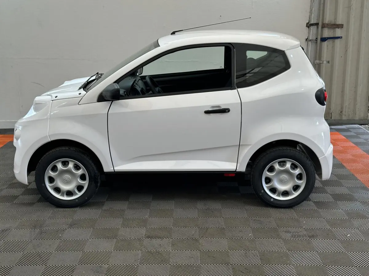 Vue de profil côté gauche d'une Aixam Minauto E Access blanche électrique avec rétroviseur caméra conducteur visible.