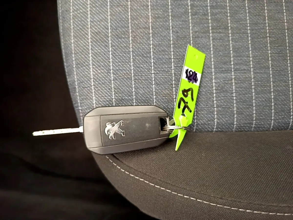 Clé de voiture Peugeot posée sur un siège tissu gris à rayures blanches, avec une étiquette verte attachée.