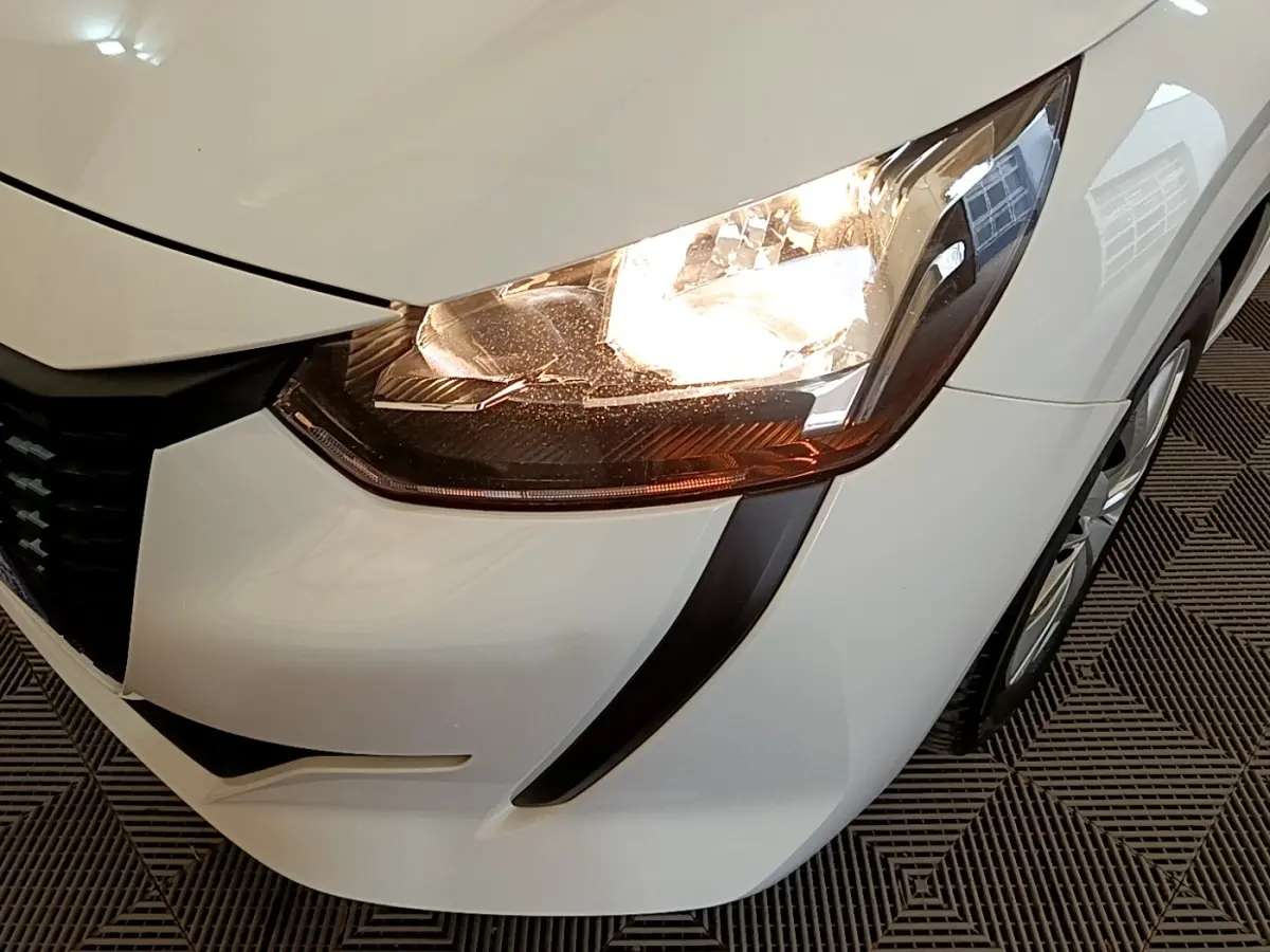 Gros plan sur l'avant droit blanc de la Peugeot 208 2021, mettant en valeur le phare allumé et la calandre noire.