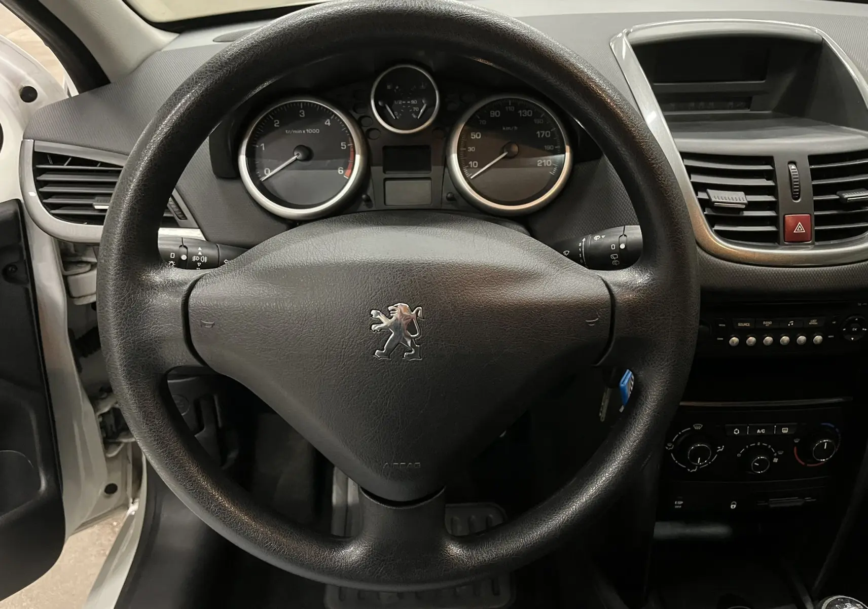 Vue intérieure centrée sur le volant noir et le tableau de bord gris de la Peugeot 207 blanche de 2011.