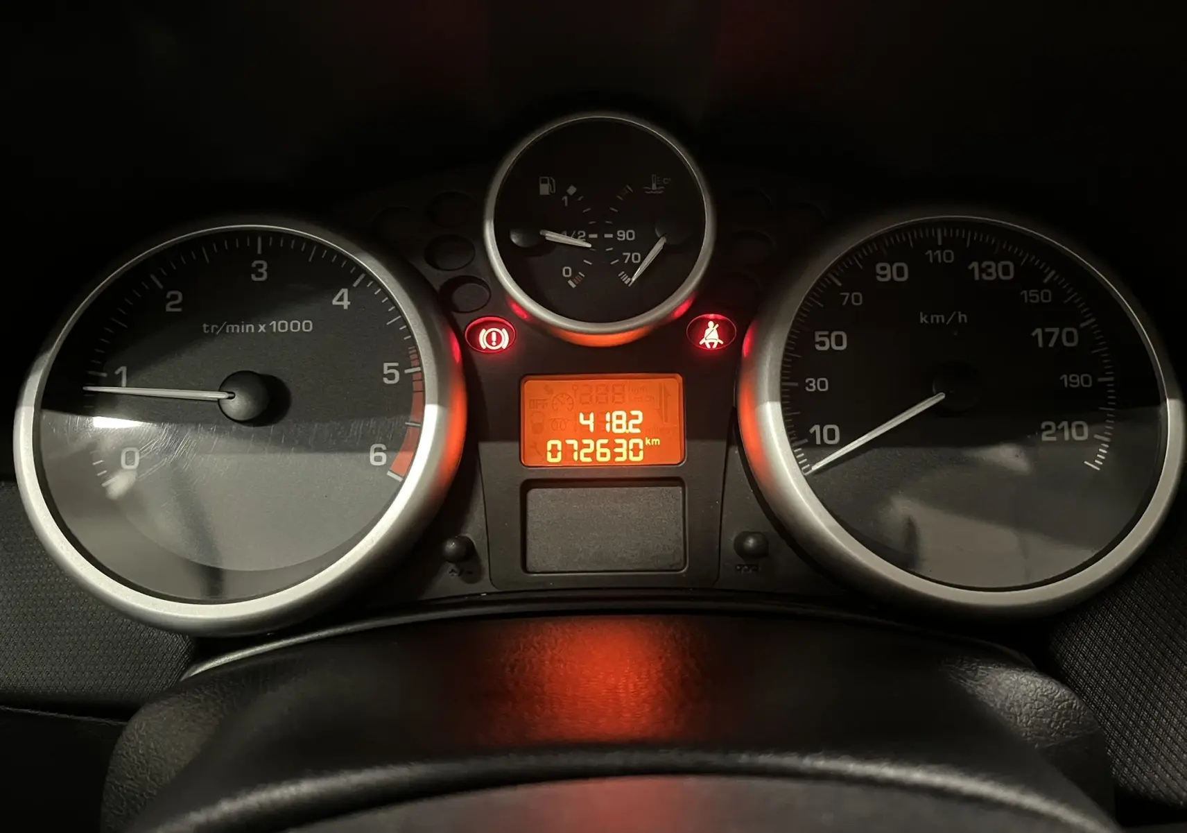 Vue rapprochée du tableau de bord de la Peugeot 207 blanche, avec compte-tours, vitesse et affichage digital orange.
