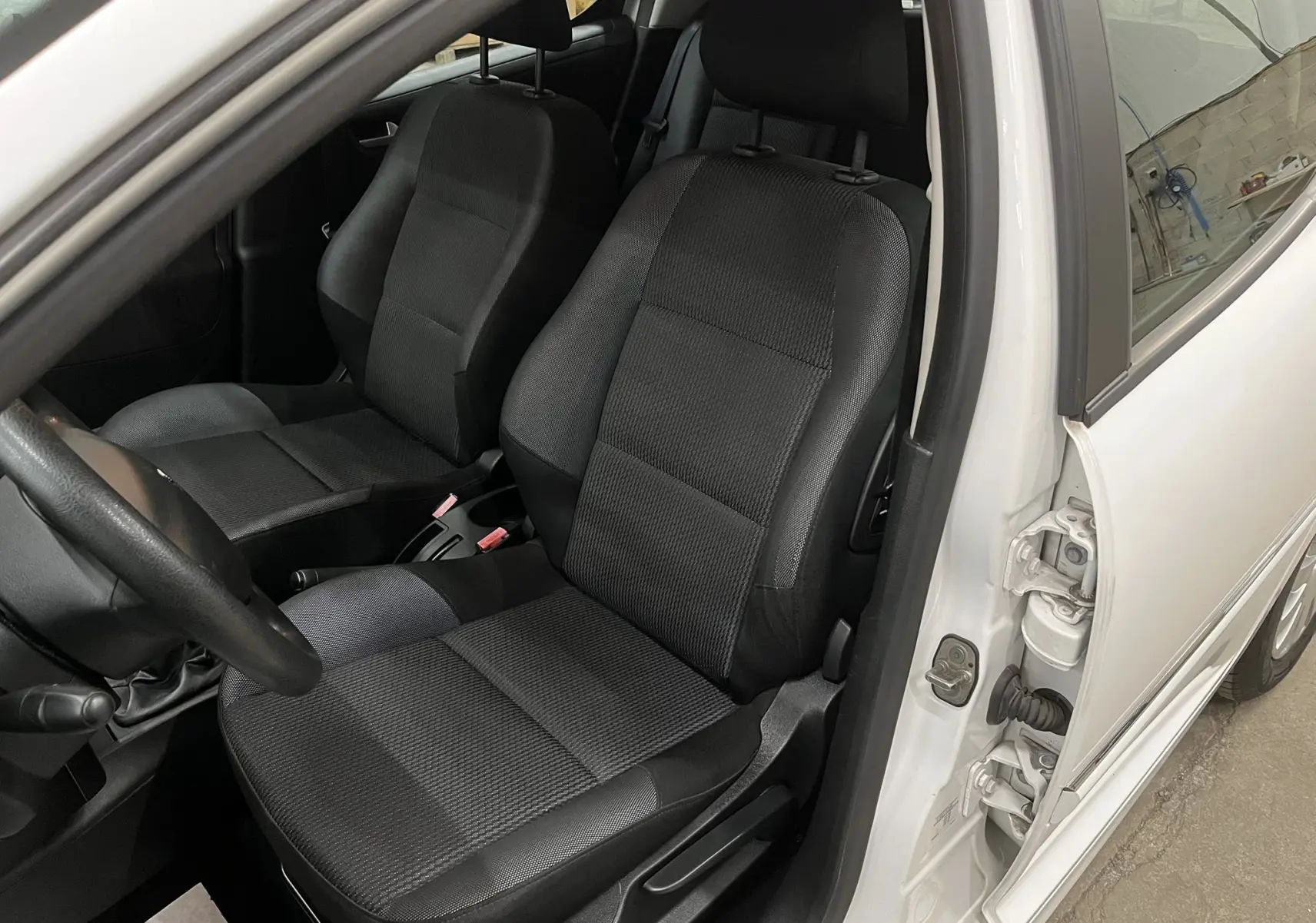 Vue intérieure côté conducteur d’une Peugeot 207 blanche de 2011, sièges tissu gris-noir et volant visible.