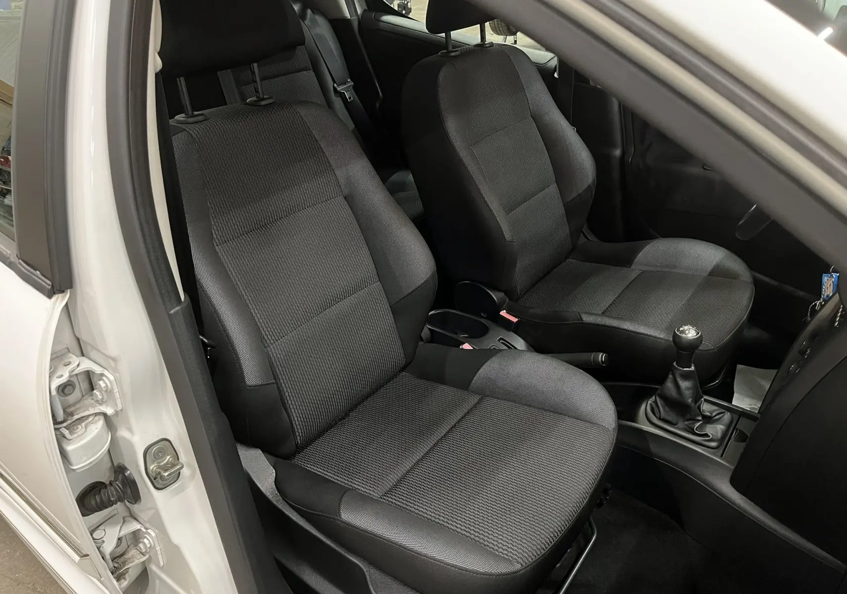 Vue intérieure côté conducteur d’une Peugeot 207 blanche, montrant les sièges avant en tissu gris noir et la boîte manuelle.