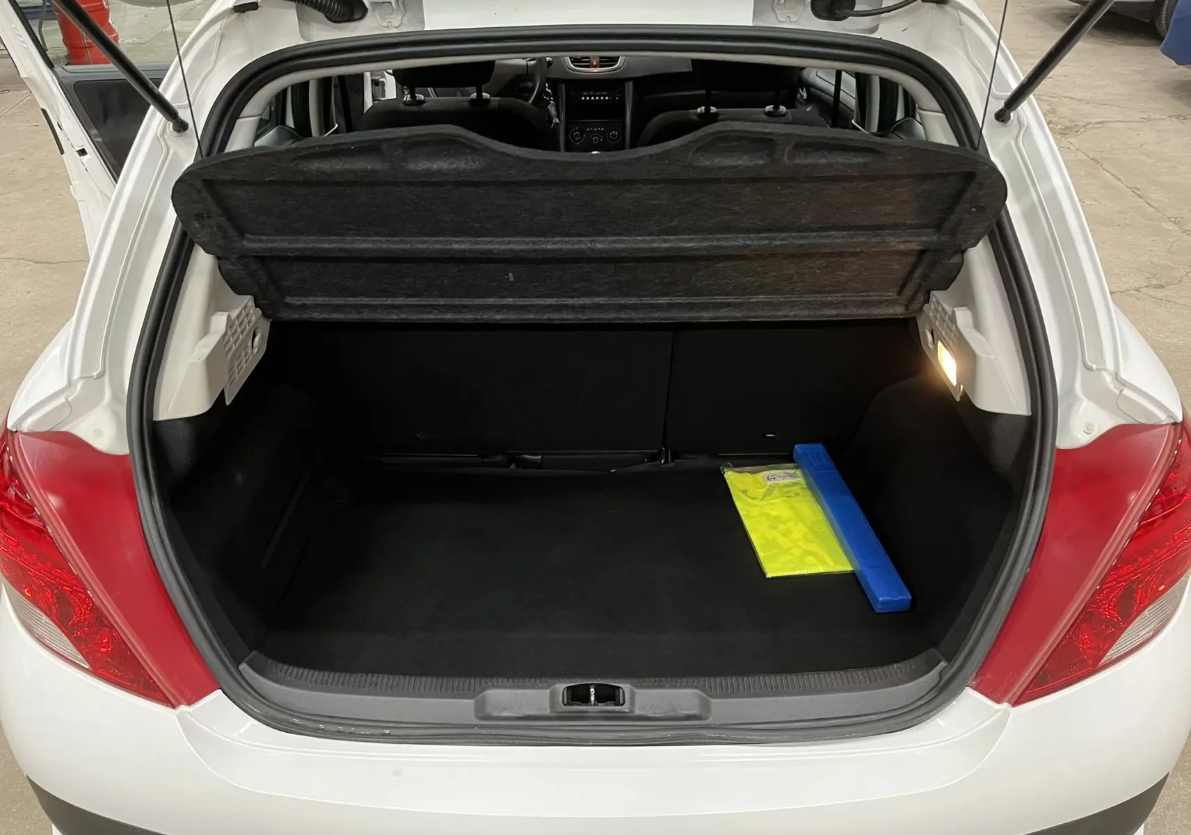 Coffre ouvert d'une Peugeot 207 blanche vue de l'arrière, avec tapis noir et gilet de sécurité visible à droite.