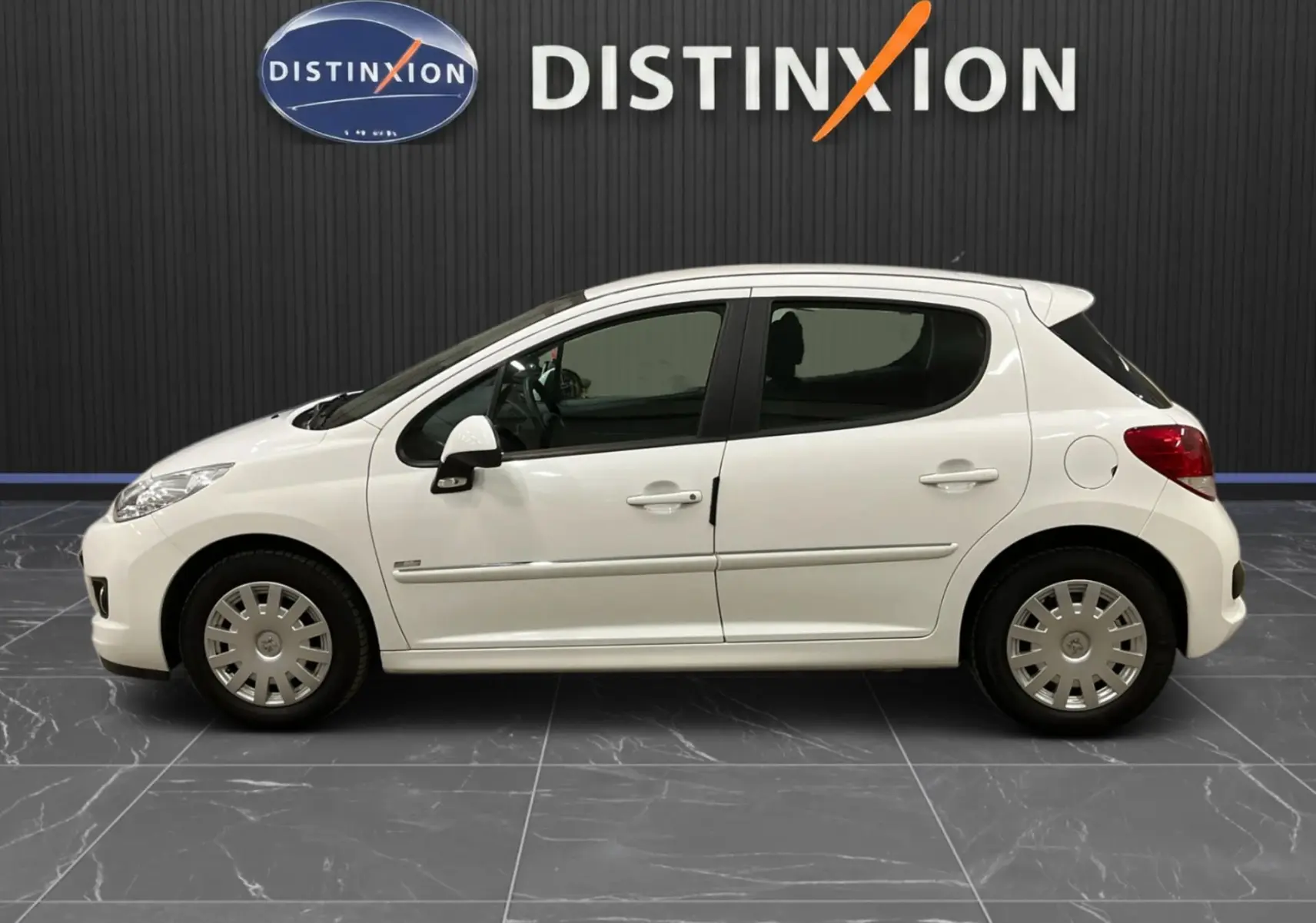 Profil droit d'une Peugeot 207 blanche 5 portes avec baguette chrome et jantes acier en intérieur showroom.