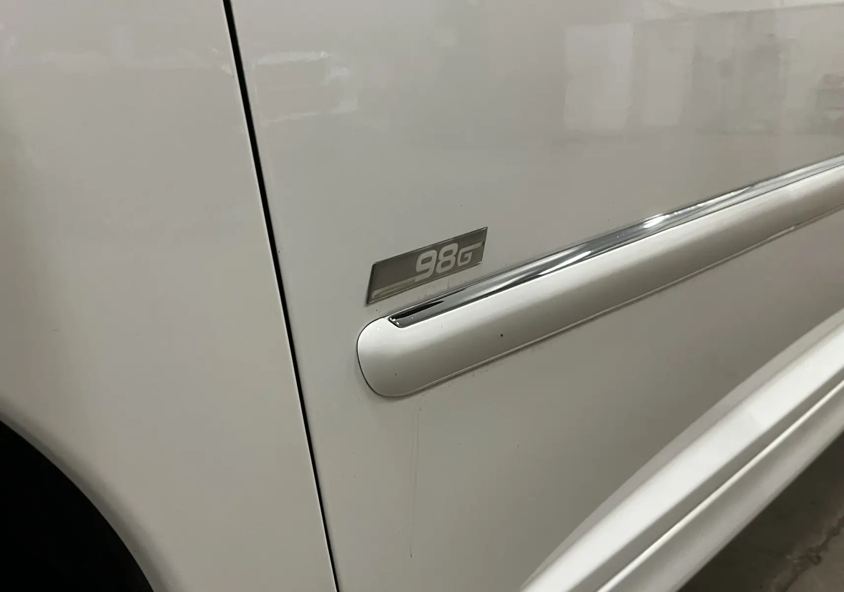 Gros plan sur la porte côté gauche d'une Peugeot 207 blanche avec badge finition 98G et baguette chromée.