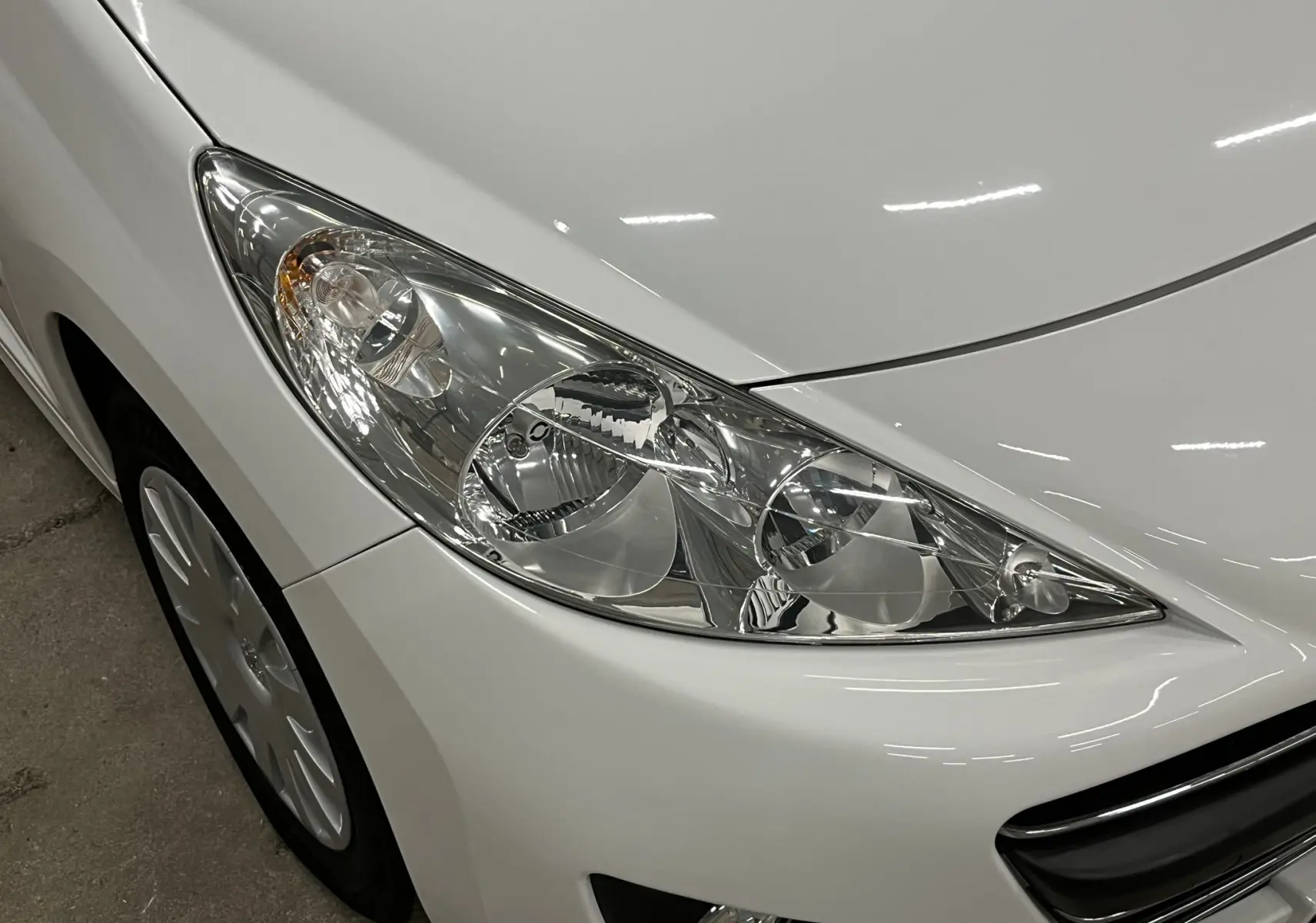 Gros plan sur le phare avant droit d'une Peugeot 207 blanche, avec calandre et jante visibles.