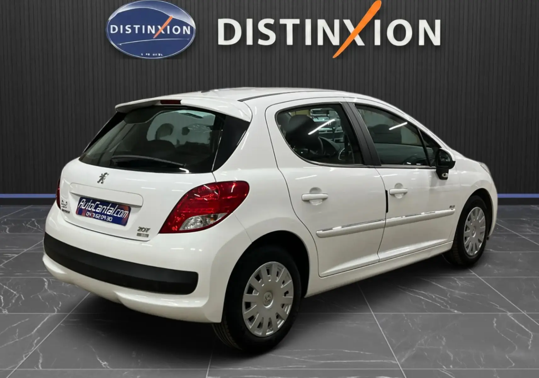 Vue 3/4 arrière droite d'une Peugeot 207 blanche finition Active avec baguettes chrome et béquet, en intérieur showroom.