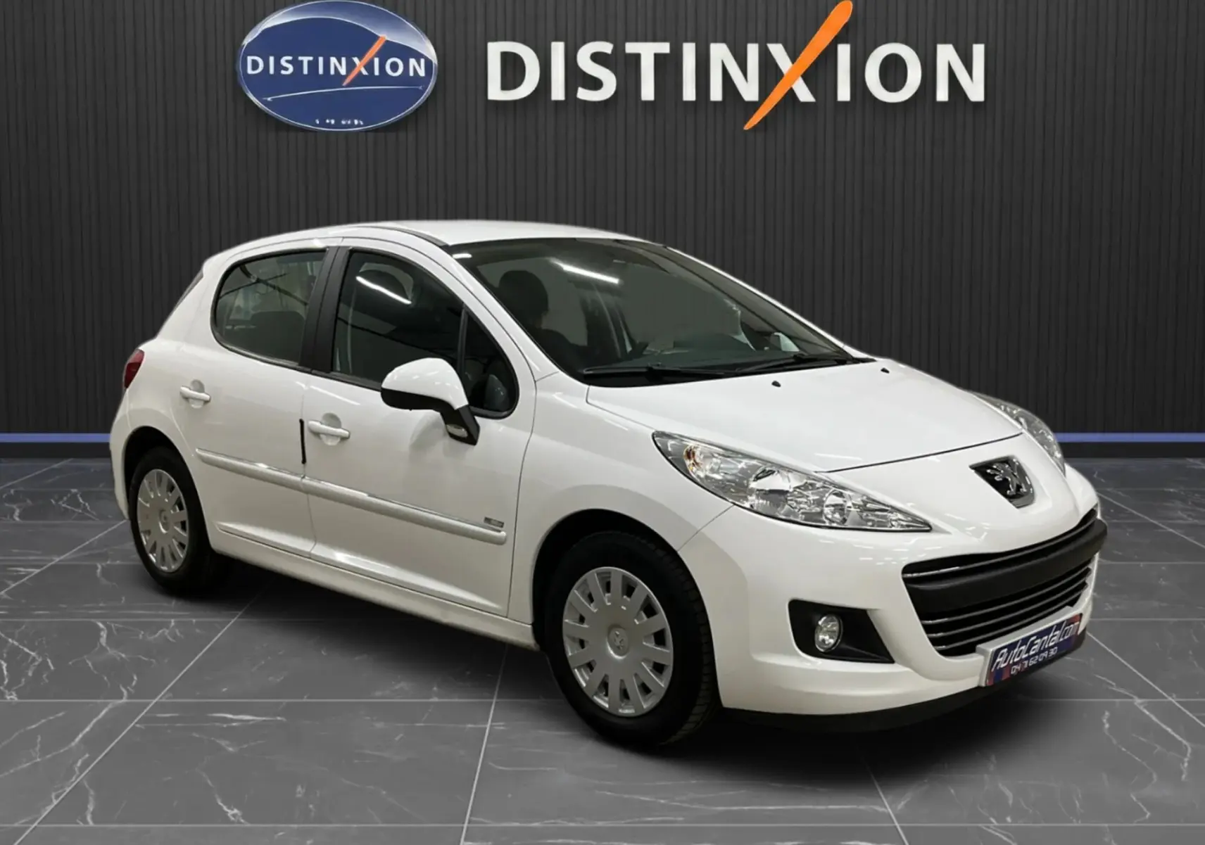Peugeot 207 blanche vue en 3/4 avant droit, avec calandre et baguettes de porte chromées, stationnée en intérieur.