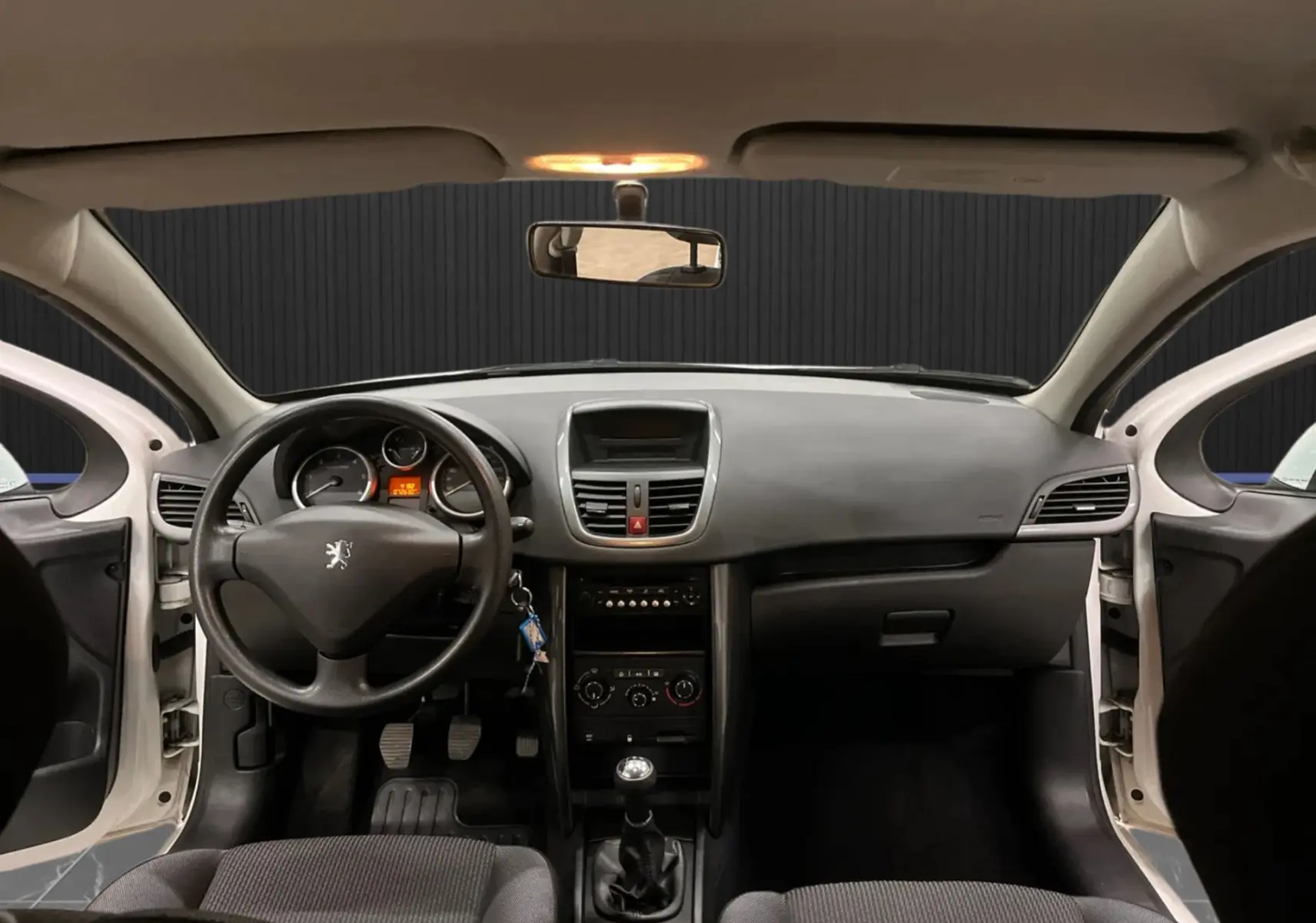Vue intérieure frontale du tableau de bord d'une Peugeot 207 blanche de 2011 avec volant, levier de vitesse manuel et sièges tissu gris-noir.