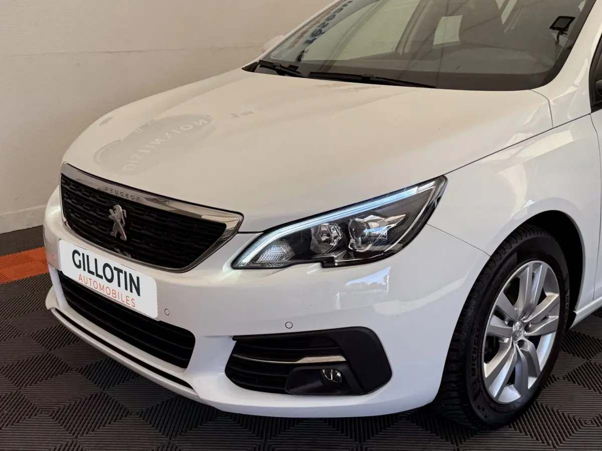 Vue 3/4 avant droit d'une Peugeot 308 blanche avec calandre noire et phares LED distinctifs.