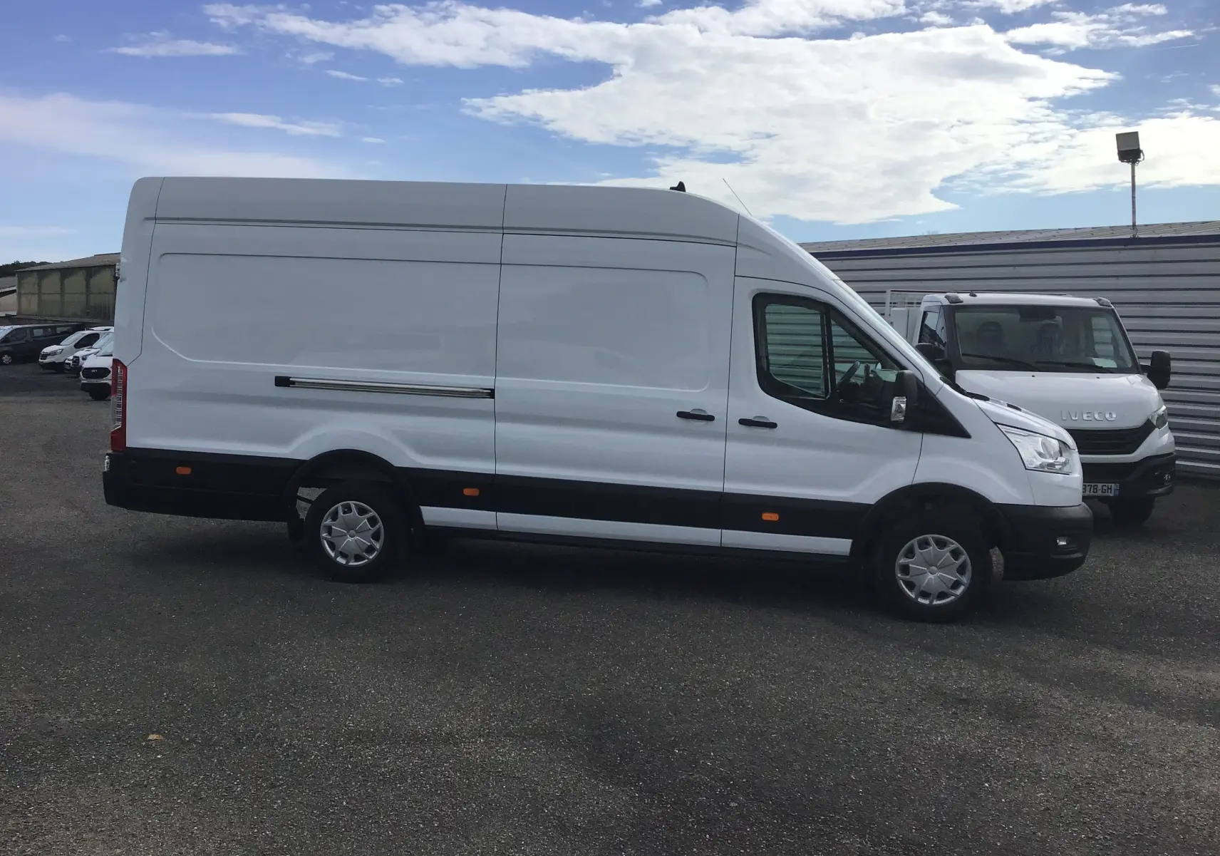 Fourgon Ford Transit blanc vu de profil côté gauche sur un parking avec un ciel partiellement nuageux.