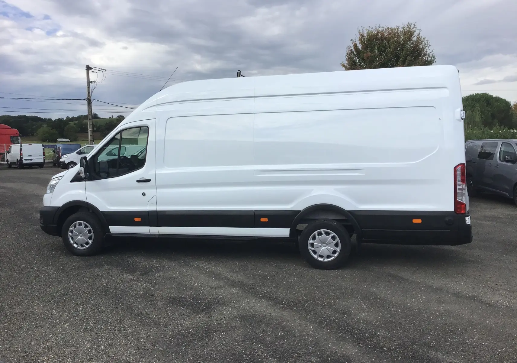 Vue latérale gauche d'un fourgon Ford Transit blanc à toit élevé avec protections noires et feux arrière verticaux.