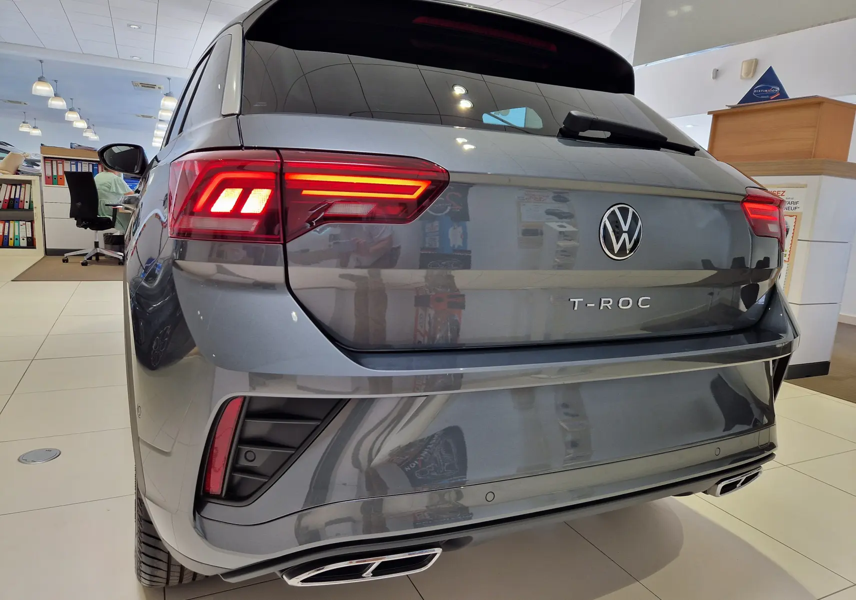 Vue 3/4 arrière du Volkswagen T-Roc gris Indium Metal 2025 avec feux LED allumés et double sortie d'échappement rectangulaire.