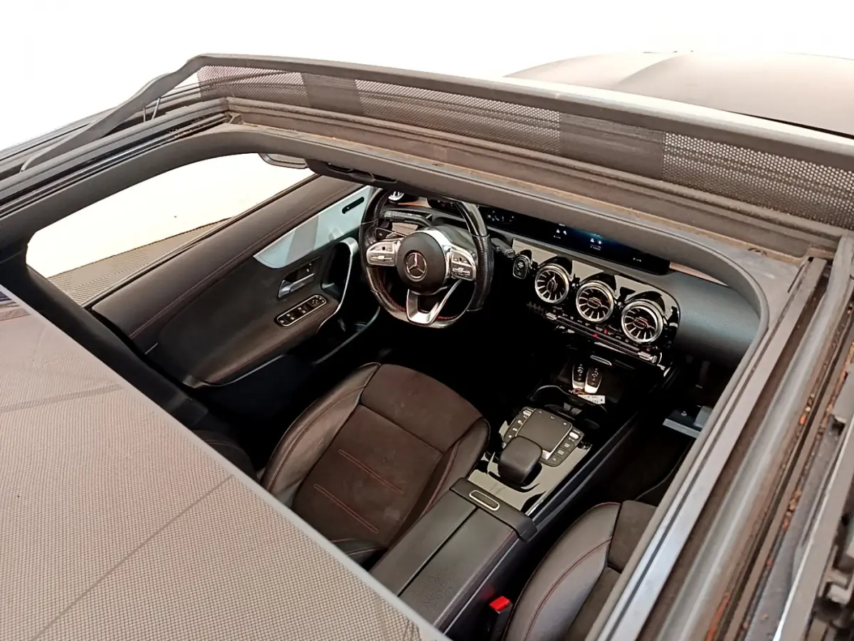 Vue intérieure par le toit ouvrant du poste de conduite noir avec volant multifonction et aérateurs ronds du Mercedes CLA 200 d AMG Line.