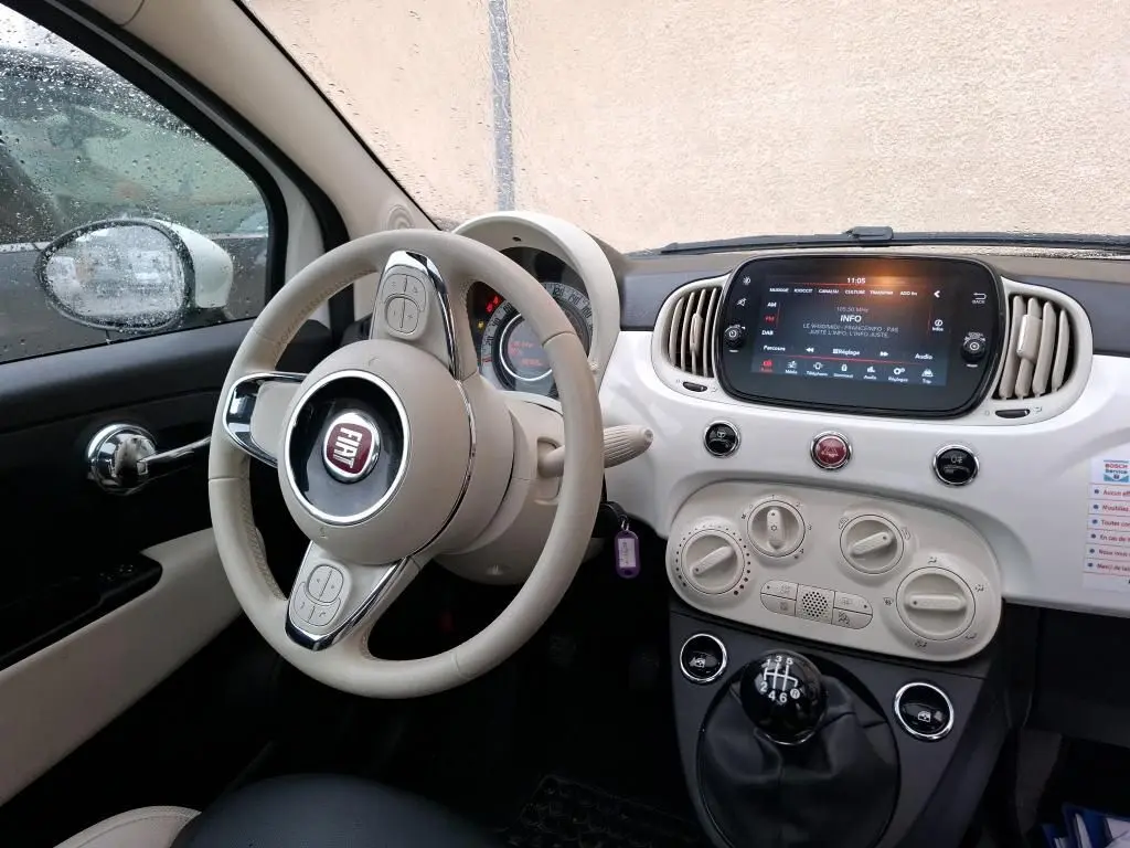Vue intérieure côté conducteur du tableau de bord blanc et beige d'une FIAT 500 2022 avec volant beige et levier de vitesses manuel noir.