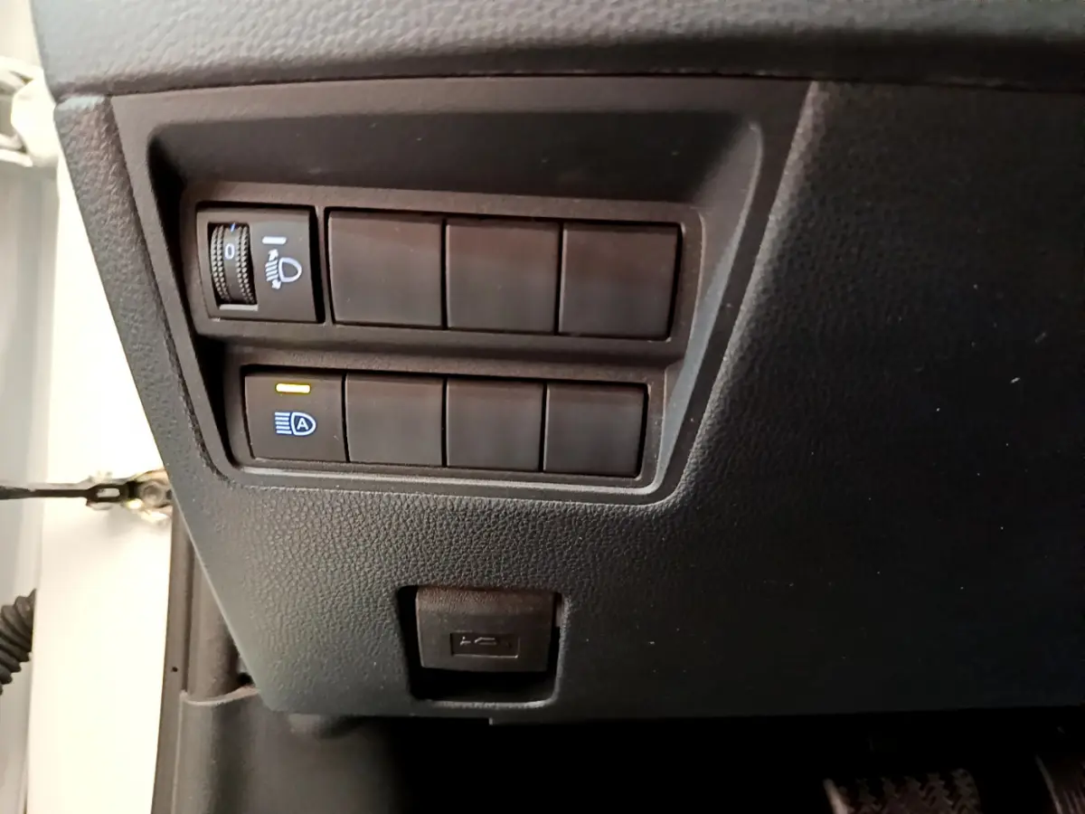Gros plan sur les commandes d'éclairage et un port USB sous le tableau de bord côté conducteur d'une Toyota Yaris Cross blanche.