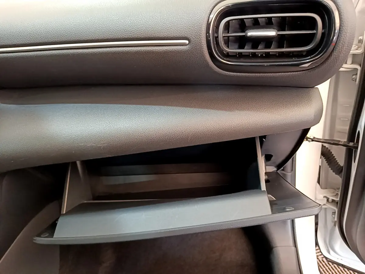 Gros plan sur la boîte à gants ouverte côté passager du Toyota Yaris Cross blanc, montrant l'intérieur noir du tableau de bord.